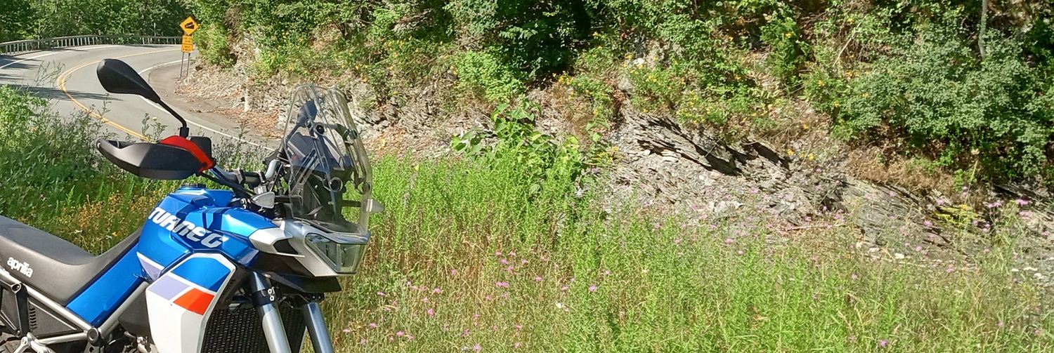 Bue, white, and red Aprilia Tuareg parked in a grassy overlook above a curve in a mountain road.