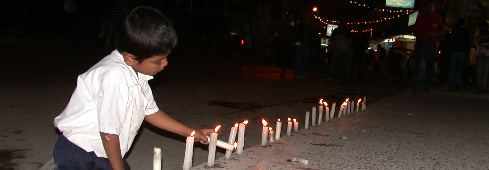 child lighting a row of candles along a dark street with colored lights overhead