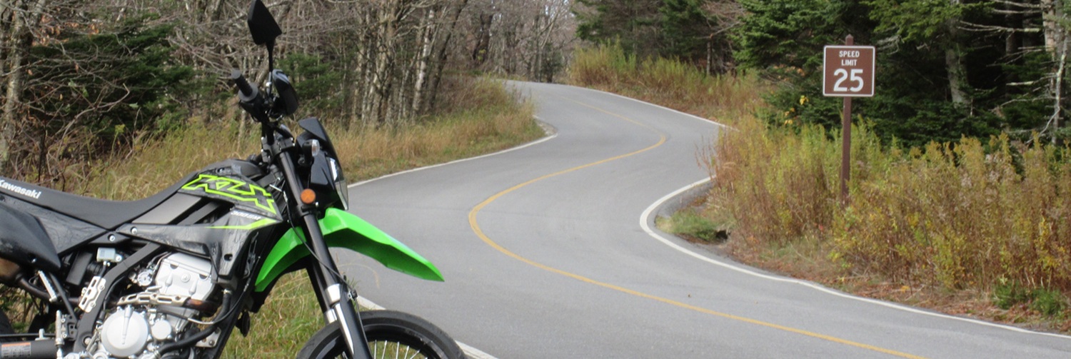 Green and black Kawasaki KLX300SM motorcycle parked along a narrow, curvy forest road with a 25 mph speed limit sign