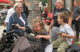 adults look on laughing at two small children smiling and sitting on a motorcycle